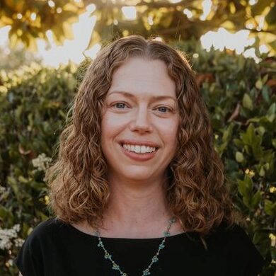 A person with curly hair smiling in front of leafy greenery, wearing a necklace and a black top.