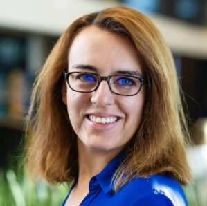 Headshot of a person with long brown hair, wearing glasses and a blue shirt, smiling in an indoor setting.