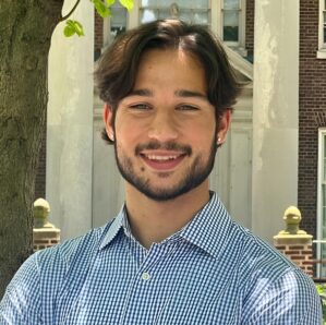 Headshot of a person with short dark hair and a beard, wearing a checkered shirt, smiling outdoors near a tree and a brick building.