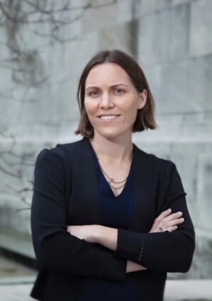A person with short hair, wearing a black blazer and blue shirt, standing with arms crossed in front of a stone wall.