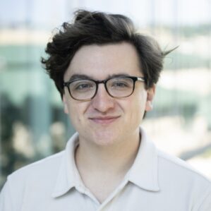 A person with short, dark hair and glasses, wearing a light-colored shirt, smiles in front of a blurred outdoor background.